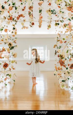 Blondes Mädchen in weißem Kleid steht ernsthaft zwischen hängenden Blumen Stockfoto