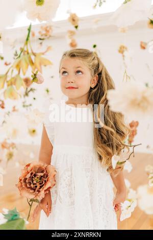 Blondes Mädchen in weißem Kleid unter Blumen schaut lächelnd auf Stockfoto