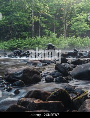 Ein ruhiger Fluss, der durch einen Wald fließt, mit einem kleinen Inukshuk, der aus gestapelten Steinen am Ufer und üppigen grünen Bäumen im Hintergrund besteht. Stockfoto