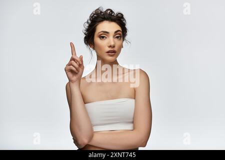 Eine junge Frau in einem weißen Oberteil zeigt ihren Finger nach oben, als würde sie eine Idee oder ein Konzept präsentieren. Stockfoto