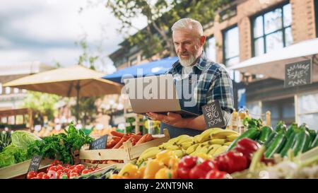 Porträt eines Straßenverkäufers mittleren Alters, der an einem Laptop arbeitet, während er auf einem Bauernmarkt mit frischen natürlichen landwirtschaftlichen Produkten steht. Geschäftsmann Kontaktiert Lieferanten Online Stockfoto