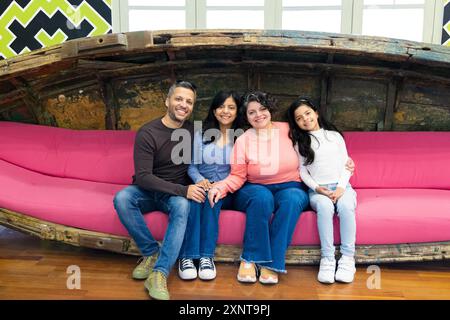 Eine vierköpfige Familie sitzt auf einer pinken Couch und posiert für ein Foto. Die Couch ist groß und rosa und die Familienmitglieder lächeln alle. Die Szene ist glücklich und W Stockfoto
