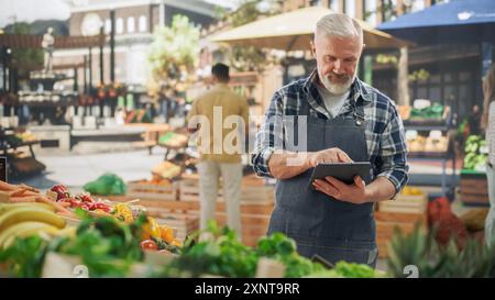 Porträt eines Straßenverkäufers mittleren Alters, der an einem Tablet-Computer arbeitet, während er auf einem Bauernmarkt mit natürlichen landwirtschaftlichen Produkten steht. Geschäftsmann, der den Lagerbestand überprüft und Lieferanten kontaktiert Stockfoto