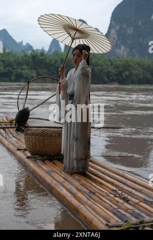 Hanfu Girl mit deng, der vom Floß auf dem Fluss Li in China aus die Berglandschaft betrachtet. Vertikal Stockfoto