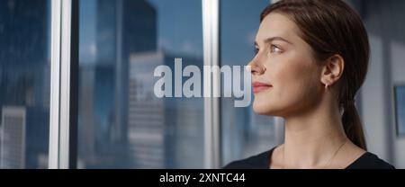 Nahaufnahme wunderschönes Porträt einer kaukasischen Geschäftsfrau, die neben dem Fenster im Big City Office mit Wolkenkratzern posiert. CEO lächelt, sieht Kamera an. Erfolgreicher Weiblicher Business Manager. Stockfoto