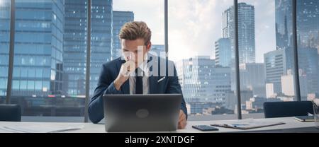 Selbstbewusster Geschäftsmann im Anzug, der an einem Schreibtisch in einem modernen Büro sitzt und einen Laptop benutzt, neben einem Fenster mit Blick auf die Stadt. Erfolgreicher Finanzmanager, Der Über Arbeitsprojekte Nachdenkt. Stockfoto