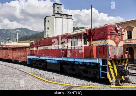 Alte rot-blaue Zugmaschine, die an einer historischen Eisenbahn mit Industriegebäuden und malerischen Hügeln im Hintergrund unter einem bewölkten Himmel stationiert ist. Stockfoto