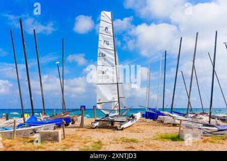 Tarragona, Spanien - 14. Juli 2024: Katamaran mit markiertem Segel am Sandstrand, umgeben von anderen Segelbooten, vor dem Hintergrund des blauen Himmels Stockfoto