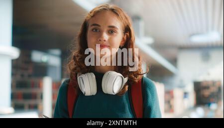 Junge Schülerin Mit Kopfhörern Um Den Hals, Lächelnd Im Modernen Schulflur. Kaukasischer Teenager mit Büchern, legeren Teal Pullover und rotem Rucksack, bereit für den Unterricht. Stockfoto