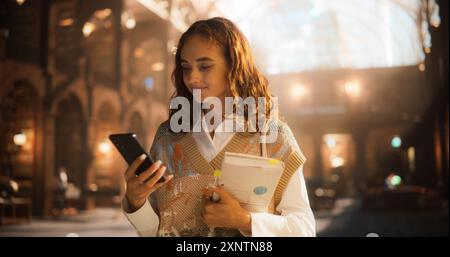 Junge kaukasische Studentin, die ihr Smartphone überprüft, während sie Bücher in einer historischen Bibliothek hält. Elegante Frau im legeren Outfit, die sich mit akademischen Aktivitäten beschäftigt, umgeben von klassischer Architektur. Stockfoto