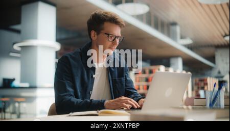 Konzentrierter junger kaukasischer männlicher Student, der an seinem Laptop in einer modernen Bibliothek arbeitet. Mann beschäftigte sich mit akademischer Forschung mit Books Around, fokussiert auf Bildschirm. Stockfoto
