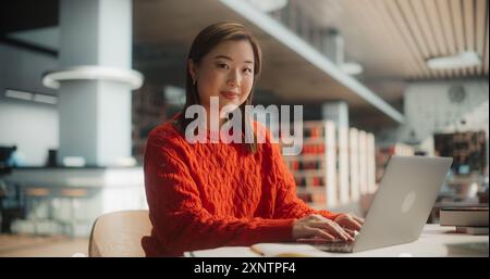Selbstbewusste asiatische Studentin, die an einem Laptop in einer modernen Bibliothek arbeitet. Junge Frau im roten Pullover, die sich mit akademischer Forschung beschäftigt, umgeben von Büchern, lächelnd Kamera an. Stockfoto