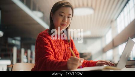 Konzentrierte asiatische Studentin, die an einem Projekt mit ihrem Laptop in einer modernen Bibliothek arbeitet. Junge Frau schreibt Notizen, engagiert in akademischer Forschung und Studienvorbereitung. Stockfoto