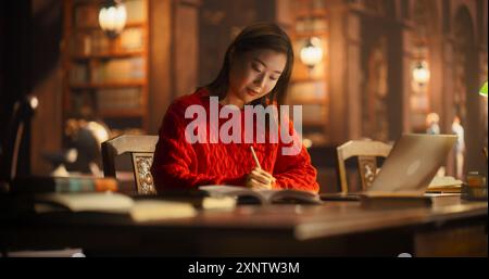 Fleißige asiatische Studentin beschäftigte sich mit akademischer Forschung in einer gemütlichen Bibliothek, schrieb Notizen und benutzte einen Laptop für E-Learning. Junge Frau in Red Sweater konzentriert sich auf das Lernen. Stockfoto