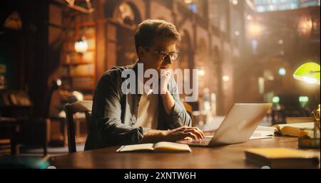 Konzentrierter junger kaukasischer männlicher Student, der sich mit E-Learning mit Laptop in einer Vintage-Bibliothek beschäftigt, umgeben von Büchern und warmer Umgebungsbeleuchtung Stockfoto