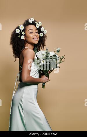 Eine schöne afroamerikanische Frau in einem weißen Kleid, mit Blumen geschmückt, hält einen Blumenstrauß und lächelt strahlend vor einem beigen Hintergrund. Stockfoto