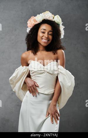 Eine afroamerikanische Braut posiert anmutig in einem fließenden weißen Hochzeitskleid, das mit einer Blumenkrone vor grauem Hintergrund verziert ist. Stockfoto