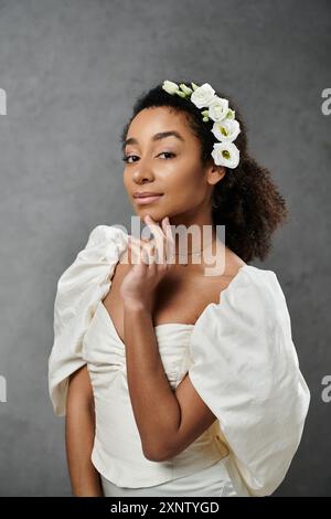 Eine wunderschöne afroamerikanische Braut, die mit weißen Blumen geschmückt ist, posiert anmutig in einem weißen Hochzeitskleid vor grauem Hintergrund. Stockfoto