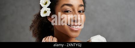 Eine wunderschöne afroamerikanische Braut lächelt hell, mit Blumen im Haar geschmückt, vor grauem Hintergrund. Stockfoto