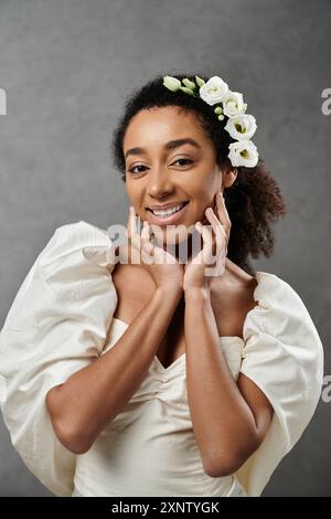 Eine wunderschöne afroamerikanische Braut in einem weißen Hochzeitskleid mit Blumen im Haar lächelt strahlend vor grauem Hintergrund. Stockfoto