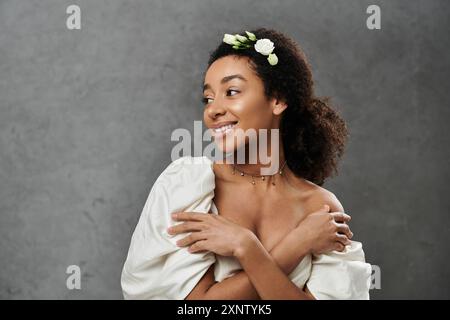 Eine afroamerikanische Braut in weißem Kleid lächelt elegant, mit weißen Blüten geschmückt, vor grauem Hintergrund. Stockfoto