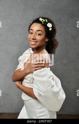 Eine wunderschöne afroamerikanische Braut in einem weißen Hochzeitskleid, ihr Haar mit Blumen verziert, lächelt selbstbewusst vor grauem Hintergrund. Stockfoto