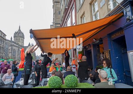 Royal Mile, Edinburgh, Schottland, Großbritannien. August 2024. Erster Tag des Edinburgh Festival Fringe-Darsteller unterhalten sich auf einer windigen Royal Mile. Pictud: Alle Hände an Deck für das Personal in Gordon's Trattoria, um die Markise an einem windigen Nachmittag zu nehmen. Quelle: Arch White/Alamy Live News. Stockfoto