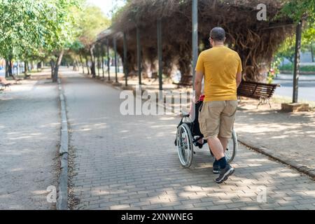 Mann auf dem Rücken schiebt einen Rollstuhl mit einer älteren Frau, die draußen in einem öffentlichen Park sitzt. Stockfoto
