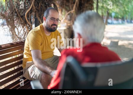 Junger Mann, der auf einer Bank sitzt und glücklich mit seiner weißhaarigen Großmutter im Rollstuhl redet. Stockfoto