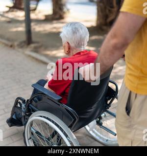 Alte Frau mit weißem Haar, die im Rollstuhl fährt und einen jungen Mann schiebt. Stockfoto