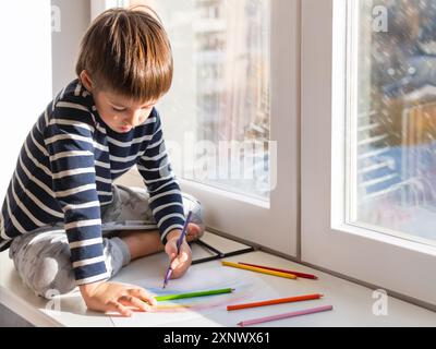 Linkshänder sitzen auf der Fensterbank und zeichnen Regenbogen mit farbigen Bleistiften. Kreative Freizeitaktivitäten für Kinder zu Hause. Stockfoto