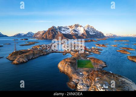 Sonnenuntergang über dem kleinen Dorf Henningsvaer, umgeben von einem schneebedeckten Massiv mit dem berühmten Fußballfeld im Vordergrund, Henningsvaer, Vagan Gemeinde, Nordland, Lofoten Inseln Stockfoto