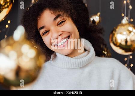 Junge Afrikanerin mit lockigen Haaren, die lächelnd zwischen goldenen Weihnachtsdekorationen steht. Lachendes schwarzes Mädchen in kuscheliger weißer Pullover. Die Dame ist genial Stockfoto