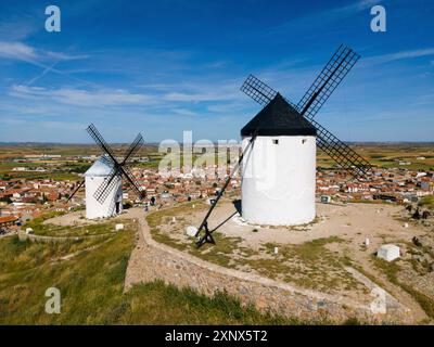 Zwei große weiße Windmühlen überblicken ein kleines Dorf in einer weiten offenen Landschaft unter einem klaren blauen Himmel, aus der Luft, Consuegra, Toledo, Castilla-La Stockfoto