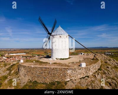 Weiße Windmühle auf einem steinernen Hügel mit Blick auf ein Dorf und Felder unter blauem Himmel, aus der Vogelperspektive, Consuegra, Toledo, Castilla-La Mancha, Spanien Stockfoto