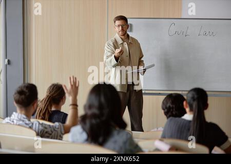 Mittlere Vollaufnahme eines erwachsenen männlichen College-Professors, der einen Vortrag über Recht im Universitätsklassenzimmer hält, während der Student die Hand hebt und Fragen stellt Stockfoto