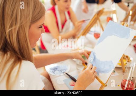 Nahaufnahme der Leinwand an Staffelei und Hand mit Spachtel. Der junge Künstler malt am Tisch ein Bild. Blondine in der Malwerkstatt. Mädchen lernt Zeichnen in der Kunst Stockfoto