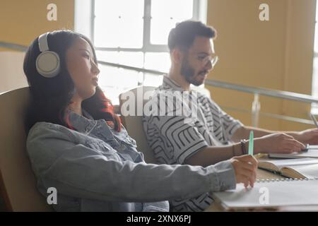 Seitenansicht einer müden asiatischen Studentin in Kopfhörern am Schreibtisch neben dem Fenster, die an einem Vortrag teilnimmt oder einen Essay mit Musik an der Universität schreibt Stockfoto