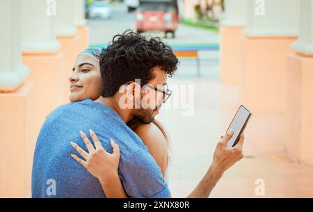 Untreuer Mann, der auf das Handy schaut, während er seine Freundin im Freien umarmt. Untreuer Freund umarmt seine Freundin und schaut auf die Zelle Stockfoto