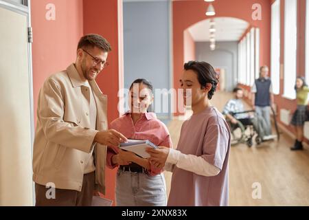Zwei glückliche Studenten sprechen beiläufig mit einem lächelnden, erwachsenen männlichen Professor und fragen ihn um Rat im pinken Universitätsflur, Kopierraum Stockfoto