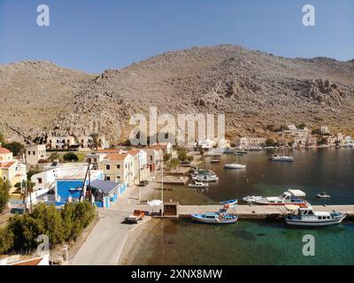 Vogelperspektive auf die Küste und die Hügel der Pedi Bay auf Symi Island, Dodekanese, Griechenland. Es gibt bunte Gebäude, Boote und einen Bootssteg. Stockfoto