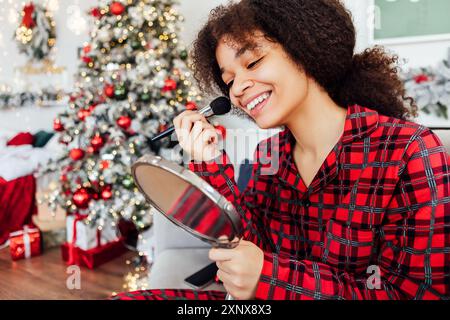 Niedlicher Afrikaner im Pyjama, der in den Spiegel schaut und festliches Make-up macht. Ein lächelndes schwarzes Mädchen trägt mit dem Pinsel Rouge auf die Wangen auf. Die Frau wird wieder Stockfoto