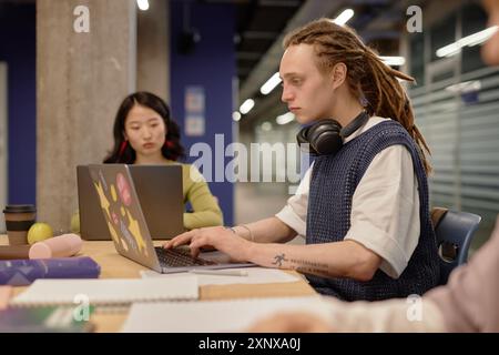 Seitenansicht eines jungen, nicht-konformen männlichen Studenten mit Dreadlock, der Schreibfähigkeiten übt und auf Laptop studiert, während er am großen Schreibtisch des Universitätsgruppenseminars sitzt Stockfoto