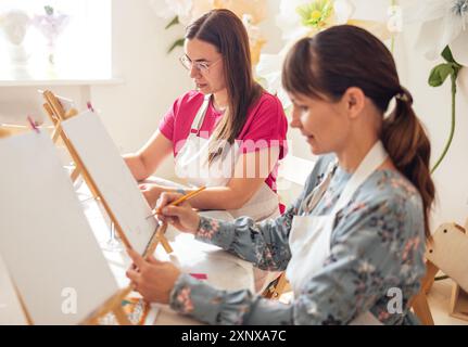 Nahaufnahme junger Frauen in Freizeitkleidung in einer Malwerkstatt. Freunde an einem Tisch mit Staffeleien und leeren Leinwänden. Die Aussicht von der Seite. Mädchen lea Stockfoto