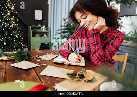 Fröhliche junge Afrikanerin im karierten Pyjama sitzt am Tisch und unterzeichnet Neujahrsgrußkarten. Das süße schwarze Mädchen wickelt zu Hause Geschenke ein. Gemütliches ich Stockfoto