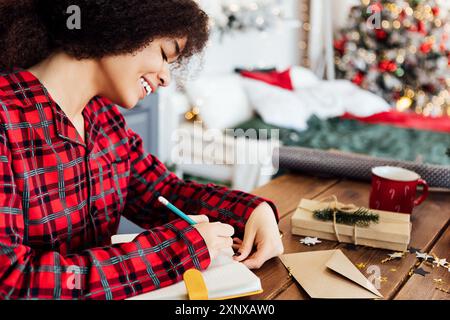 Fröhliche junge Afrikanerin im karierten Pyjama sitzt am Tisch und unterzeichnet Neujahrsgrußkarten. Das süße schwarze Mädchen wickelt zu Hause Geschenke ein. Gemütliches ich Stockfoto