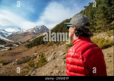 Ein langhaariger junger Mann in roter Daunenjacke und grauer Sonnenbrille steht mit dem Rücken zur Kamera und schaut auf eine Bergschlucht. Mit Stockfoto