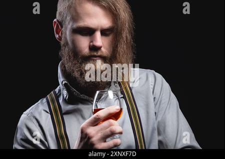 Ein attraktiver Mann mit langem Bart und Schnurrbart, der an der Wand sitzt, schnüffelt den Duft eines alkoholischen Getränks in einem Glas, das seinen hält Stockfoto