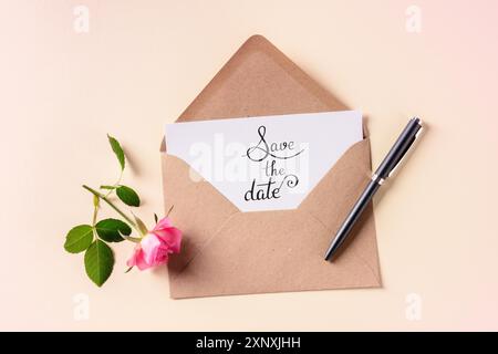 Speichern Sie den Datumsschriftzug auf einer Einladung in einem braunen Kraftumschlag, aufgenommen von oben auf einem rosa pastellfarbenen Hintergrund mit einer Rose Stockfoto