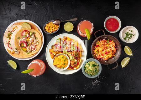 Mexikanisches Essen, eine flache Lay Overhead Aufnahme auf schwarzem Hintergrund. Nachos, Tacos, Guacamole, Garnelen, Paloma-Cocktails, Limetten und Dips Stockfoto